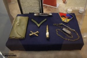 An assortment of items arranged on a dark blue display backdrop. Items include a folded pouch, pocket knife, two fabric patches, a golden pin, a bronze colored bullet and a chain necklace.