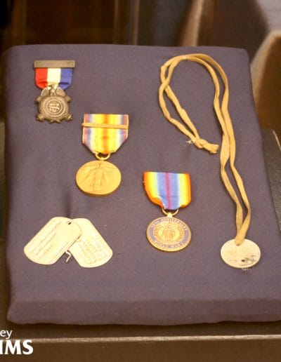 Image of five different medals and ID tags arranged on a navy blue display. Three of the medals have colorful ribbons and the ID tag on the right side has a long lanyard attached to it.