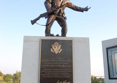 Bronze statue of a soldier wearing a uniform and helmet, carrying a gun and gesturing to his left.