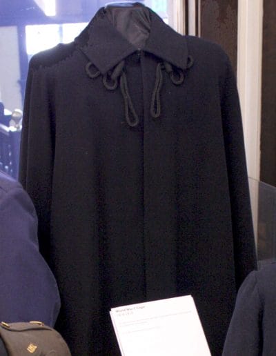 Image of a mannequin wearing a very dark blue wool cape. The cape has a collar and black decorative cords under the collar.