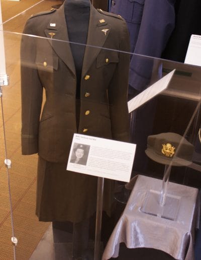 Image of a mannequin wearing a women's military uniform consisting of a brown jacket with gold pins and a brown skirt. The hat that goes with the uniform is displayed next to it.
