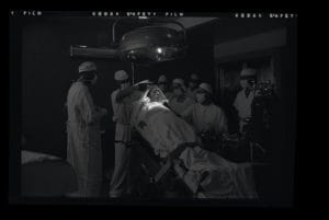 A person strapped on an operating table is spotlighted by a bright lamp on their head. Several people in surgical dress stand around behind.
