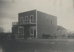 Two story storefront viewed at an angle surrounded by dirt. Farm equipment to the right and horses out front.