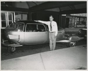 A man stands next to a three-wheeled car that has an oblong body with "wings" at the back end where the wheels extend out wider.