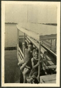 A covered dock with seating extends into a lake. Three boys sit on the railing facing the camera. 