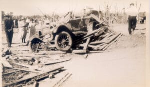 In foreground, an damaged automobile sits on a pile of debris. People mill about viewing destruction in the background.