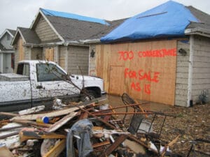 View of a heavily damaged house with boarded up windows and siding has "700 Cornerstone for sale as is" spray painted on siding.