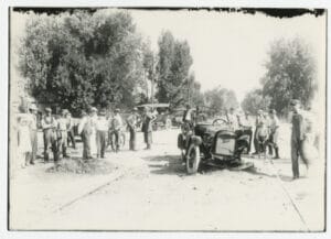 A wrecked automobile sitting on the trolley tracks surrounded by a crowd of people looking at the wreck.