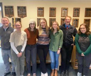 Seven adults smile for the camera against a display wall of plaques.