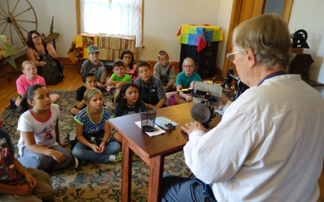 An adult sits at a table demonstrating old sewing machines to school children seated on the floor.