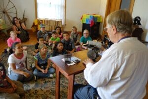 An adult sits at a table demonstrating old sewing machines to school children seated on the floor.