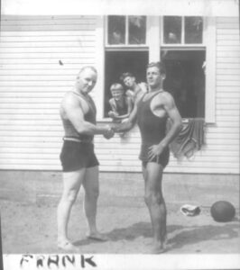 Two men shaking hands and looking at the camera, identified as (L-R) Toots Mondt and Frank Mondt. Both men are dressed in shorts and tank tops.